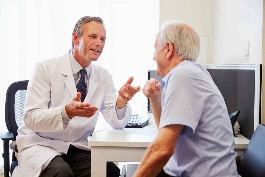 A senior man sitting in a hearing clinic office speaking to a male hearing specialist during a hearing aid consultation visit.